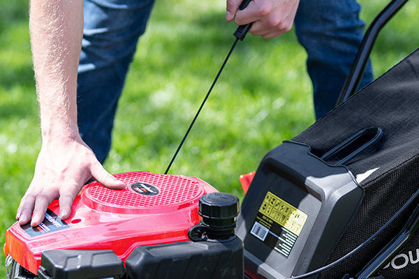 A close-up outdoor shot of a person using a red and black lawnmower, with one hand pulling the engine start cord and the other resting on the mower deck. The background shows a blurred green grass lawn.